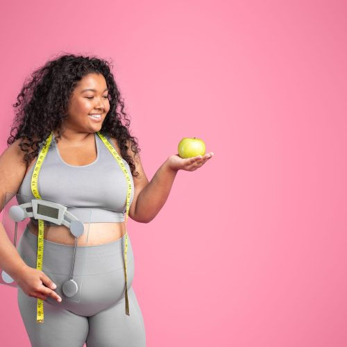 Happy african american oversize lady with measuring tape on neck holding apple and scales, isolated on pink studio background, free space. Weight loss, body care, diet and healthy food